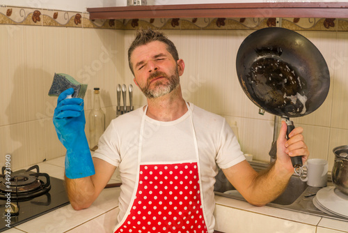 domestic chores stress - young attractive overworked and stressed home cook man in red apron hating dishwashing feeling upset and unhappy washing cooking pan photo