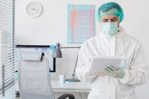 Serious medical doctor in protective workwear filling the medical card while standing at hospital