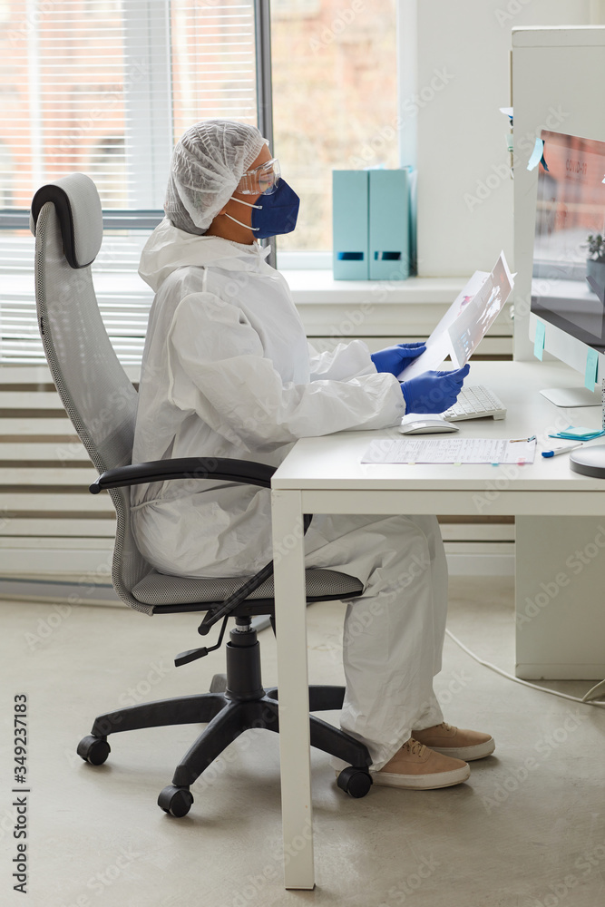 Fototapeta premium Female doctor in protective workwear sitting at the table working on computer and examining documents at office