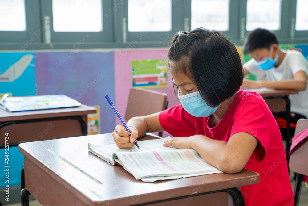 School kids wearing protective mask to Protect Against Covid-19 with pens and notebooks writing test in Classroom,Education,Elementary school,Learning and people concept. School kids wearing protective mask to Protect Against Covid-19 with pens and notebooks writing test in Classroom,Education,Elementary school,Learning and people concept.