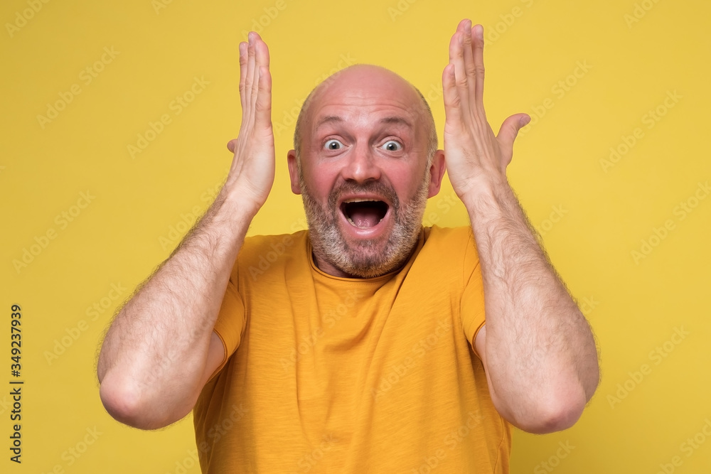 I can not believe this concept. Amazed mature caucasian man in yellow t-shirt holding hands in surprised gesture, keeping mouth wide open, looking shocked. Studio shot on yellow background.