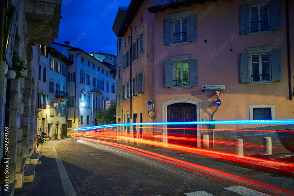 Fototapeta premium Italian cityscape evening mysterious narrow empty street by night. With car light trace. Night view of the Rovereto city