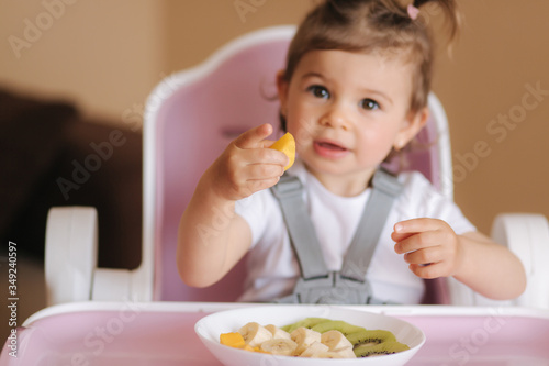 Beautiful little baby girl first time eating exotic fruits in high chair. Cute baby girl taste delitious fruist, mango, kiwi, and babana