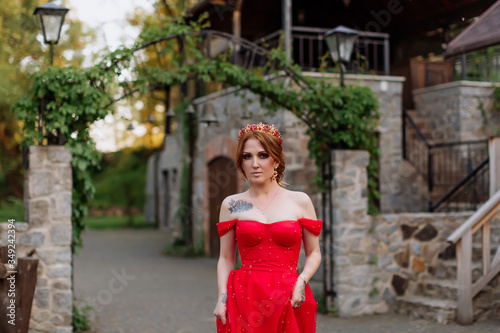 Portrait of attractive redhead tattooed woman in red dress and diadema on blurred medieval castle background © onphotoua