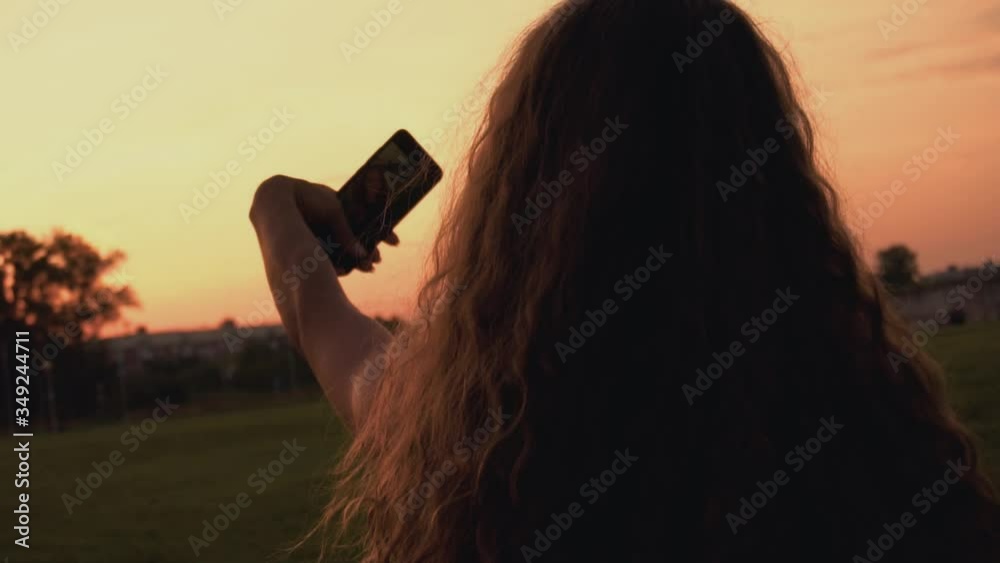 Curly long haired girl blogger takes a selfie pictures on phone at sunset for her followers or subscribers in social networking service. Rear view on back. Vogue female looks into smartphone's camera.