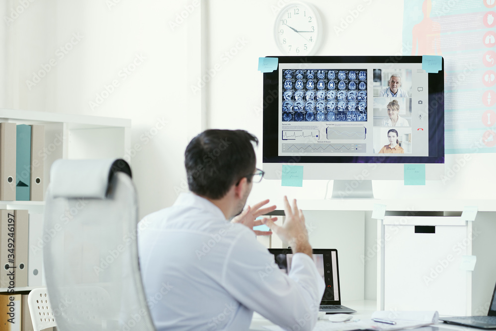 Rear view of mature male doctor sitting at the table and looking at computer monitor with x-ray images and discussing them with colleagues online