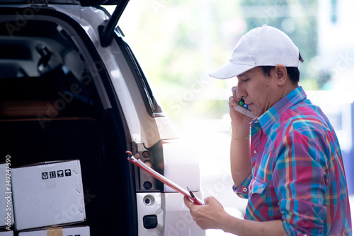 Asian delivery man calling to check the product and the delivery location, standing in back of his van, clipping board in hand