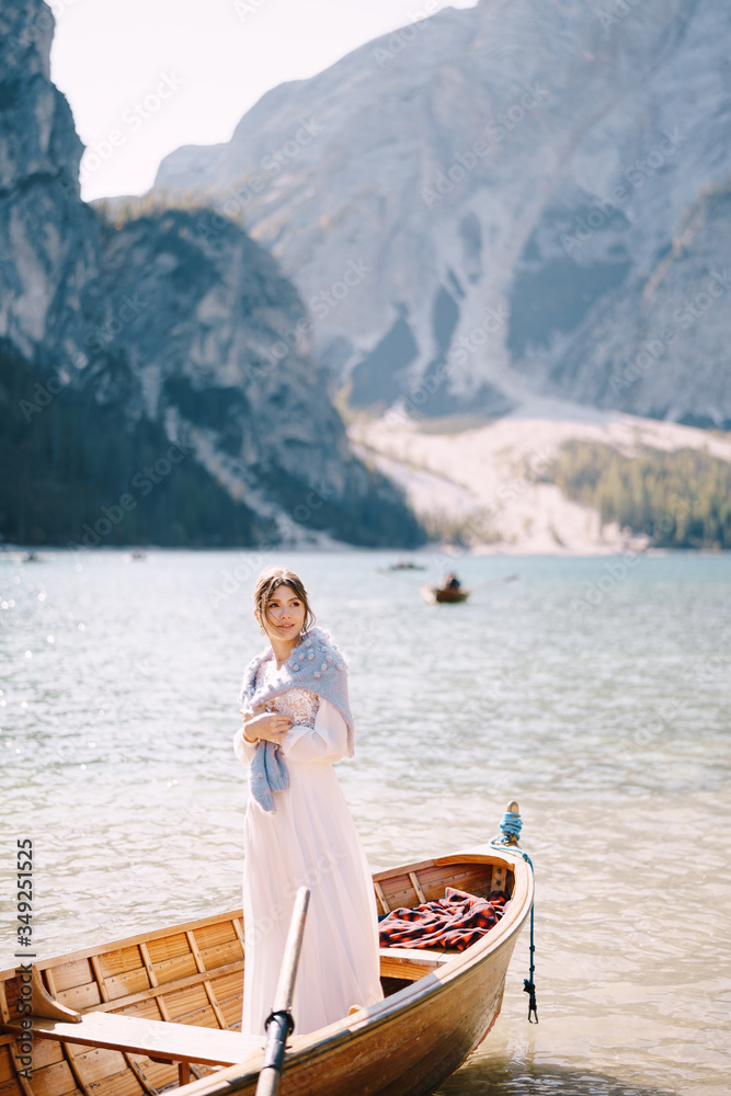 A bride stands in a wooden boat at the Lago di Braies in Italy. Wedding in Europe, on Braies lake. A young girl in a white wedding dress, covers her shoulders with a blue sweater.