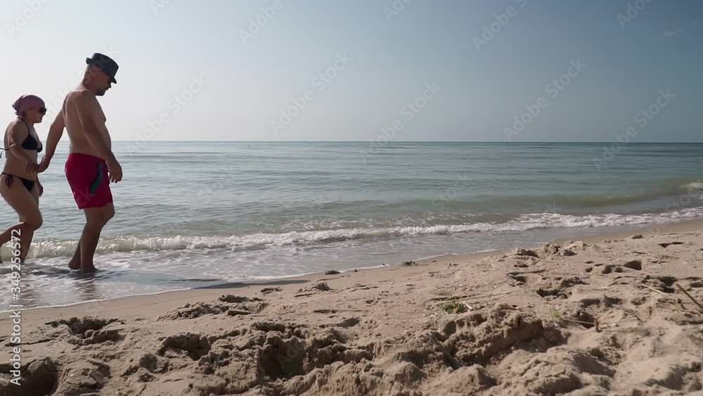 Man and woman walking along seashore on sunny day. They hold hands slowly strolling along sandy coast of Sea, irrigated by turquoise waves in warm weather. Man and woman walking along seashore on sunny day. They hold hands slowly strolling along sandy coast of Sea, irrigated by turquoise waves in warm weather.