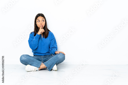 Young mixed race woman sitting on the floor isolated on white background showing a sign of silence gesture putting finger in mouth