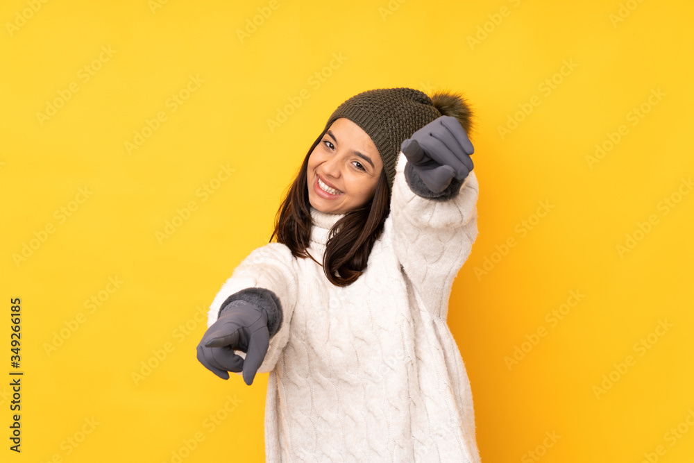Fototapeta premium Young woman with winter hat over isolated yellow background points finger at you while smiling