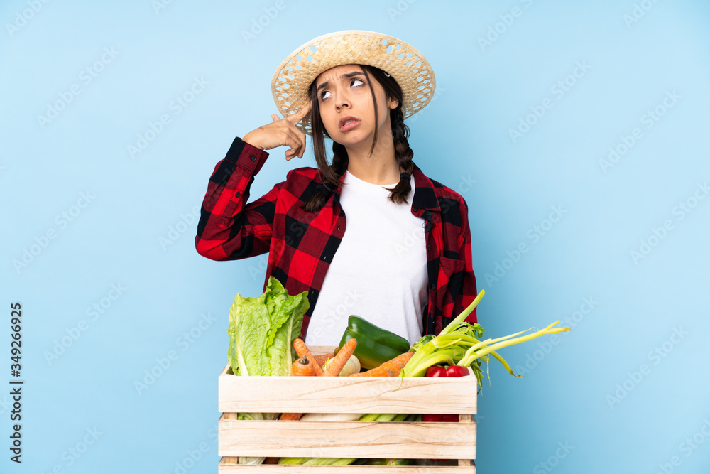 Young farmer Woman holding fresh vegetables in a wooden basket making the gesture of madness putting finger on the head