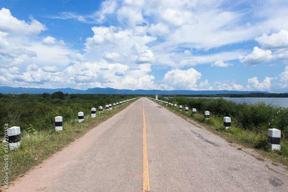 Slurry road on the ridge of the natural reservoir With mountains at the destination and clouds in the beautiful blue sky , Empty asphalt road with forests and rivers along the way. - image