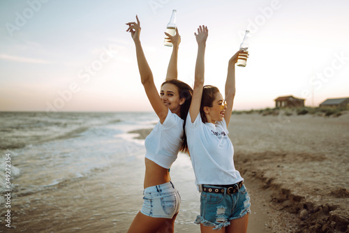 Beautiful two girls with glasses is holding a bottle with beer in her hands at the beach. Young women enjoying on beach holiday. Summer holidays, vacation, relax and lifestyle concept.