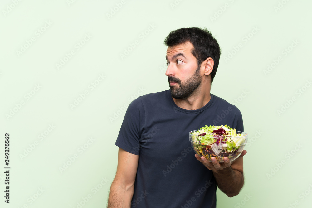 Young handsome man with salad over isolated green wall making doubts gesture looking side Young handsome man with salad over isolated green wall making doubts gesture looking side