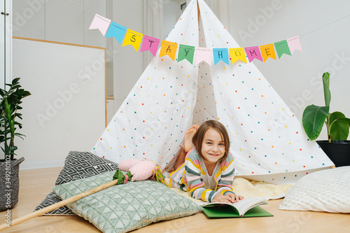 Photos 10 years old girl reading in front of a hut with stay home flag garland across