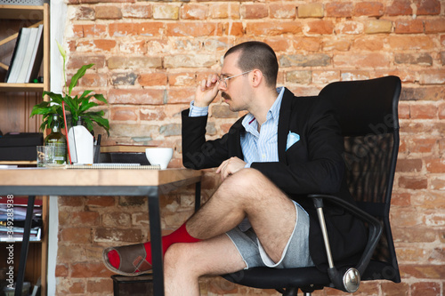 Serious. Young man without pants but in jacket working on a computer, laptop. Remote office during coronavirus, fun and comfortable in underpants. Insulation, quarantine, humor, business concept. photo