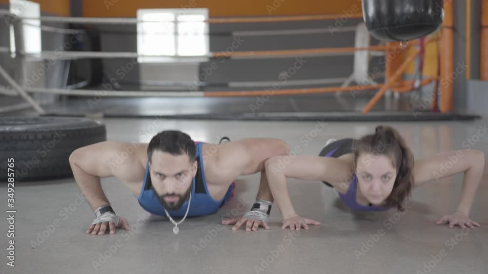 Front view of young Caucasian woman and Middle Eastern man pushing up in gym. Portrait of sportsman and sportswoman exercising in sports club together. Healthy lifestyle, endurance, sport training. Front view of young Caucasian woman and Middle Eastern man pushing up in gym. Portrait of sportsman and sportswoman exercising in sports club together. Healthy lifestyle, endurance, sport training.