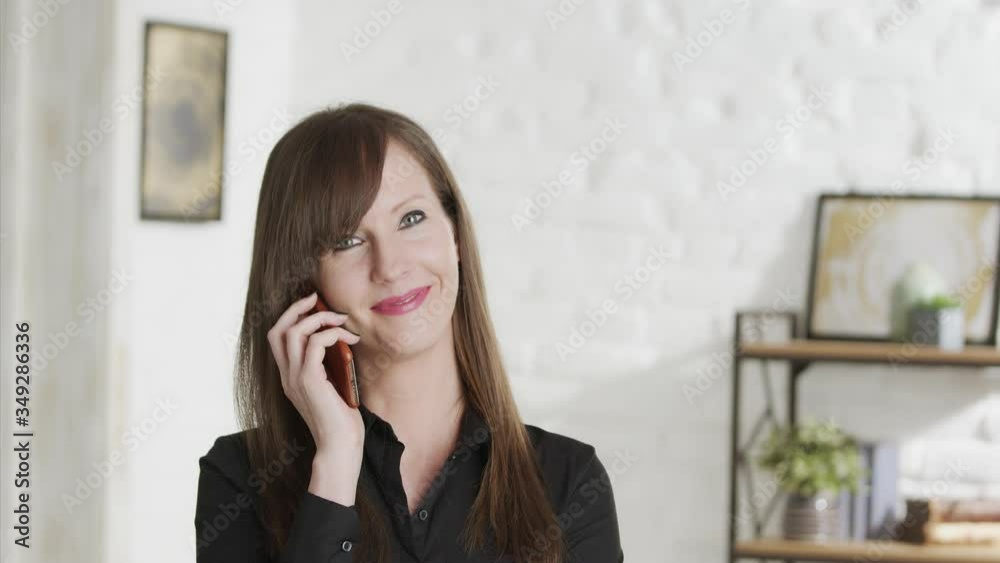 Woman in black shirt looking at camera talking on phone and smiling. Casual home in background. Woman in black shirt looking at camera talking on phone and smiling. Casual home in background.