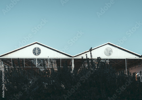 An outdoor view of two triangle metal roofs with a single round window in each one of metro or train depot buildings, outdoor storage facilities, or hangar  clear sky, dark bushes in a foreground © skyNext