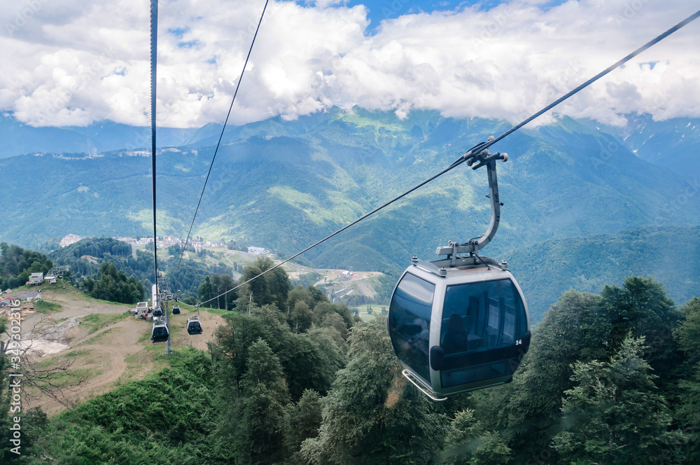 Cabins of the cableway above the tops of trees against the backdrop of mountains and clouds.