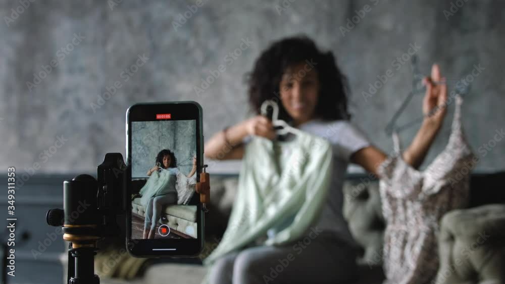 African-American fashion blogger recording video with a smartphone at home choosing clothes sitting on a sofa, rack focus, slow motion African-American fashion blogger recording video with a smartphone at home choosing clothes sitting on a sofa, rack focus, slow motion