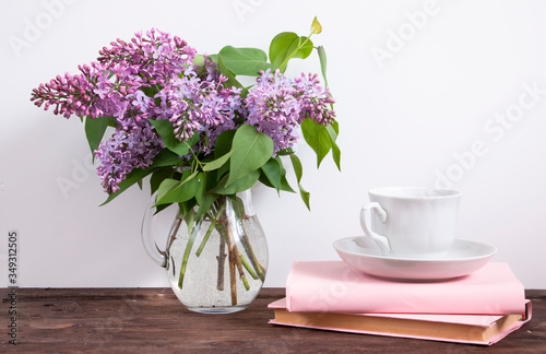 Bouquet of flowering branches of lilac on a dark wooden table. White cup and saucer and pink books. Home interior with decoration elements.