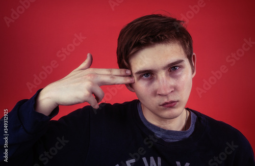 Depressed young male keeping hand gun near temple and looking at camera while pretending to commit suicide against red background