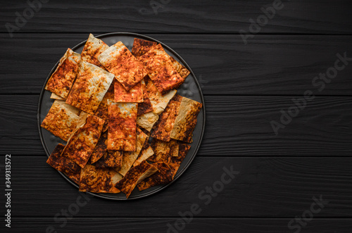 Crispy lavash nachos chips with paprika on a black plate on a black wooden table, view from above, free space