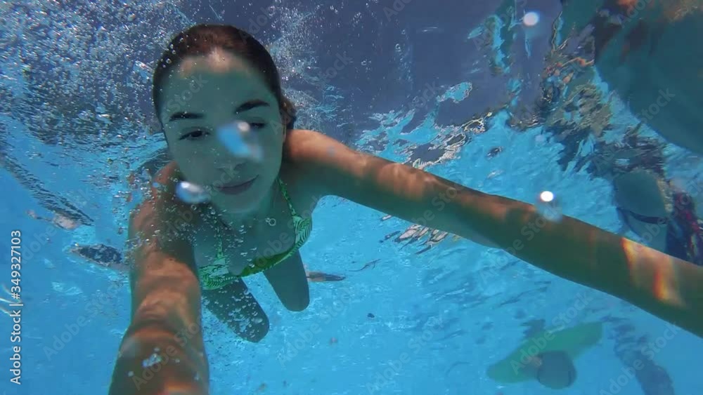 Fitted pretty woman in swimsuit diving under water in swimming pool, shooting herself on action camera. Mixed race girl filming selfie video bathing underwater. Partying with friends on beach party.