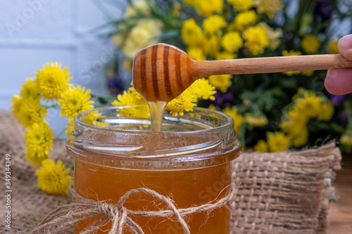 Honey jar. Dips wooden stick to glass bowl with liquid floral fresh honey. Healthy organic honey dripping or pouring from wooden spoon. Yellow background with flowers