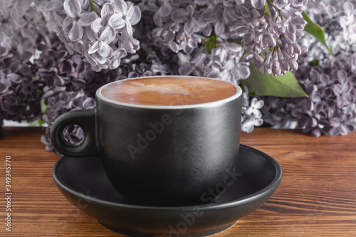 Cup of hot coffee with milk on wooden table, beautiful background with lilacs flowers