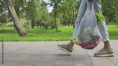 Unrecognizable woman holds cotton mesh shopping bag with vegetables and walks in the summer park