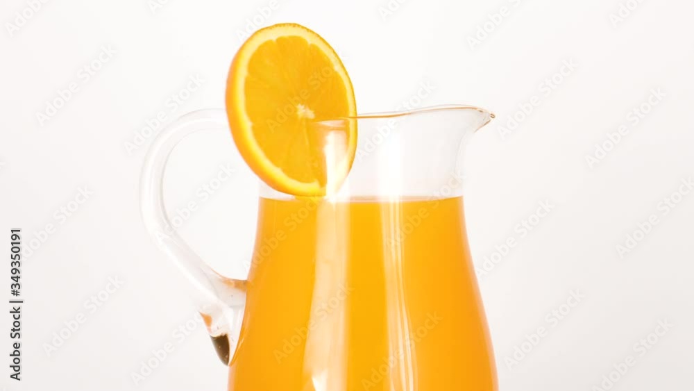 Close up of spinning jug with orange juice and slice of orange on white background Close up of spinning jug with orange juice and slice of orange on white background