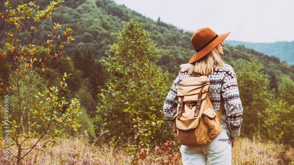 Young hipster blond hair woman in hat, jeans, wool sweater with nostalgic textile backpack looking on mountains valley. Travel, hiking and weekend activities outdoors concept.