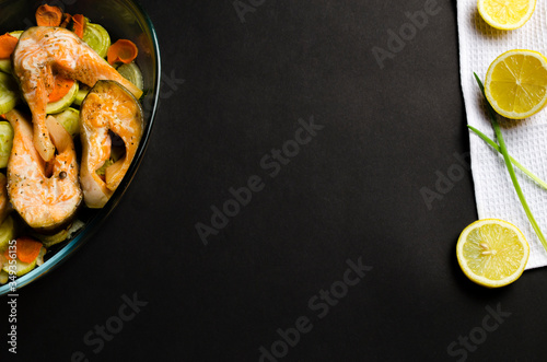 Baked salmon with vegetables: lemon, zucchini, zucchini, carrots, onions garnished with sprigs of dill in a glass tray on a black backgroundTop view. photo