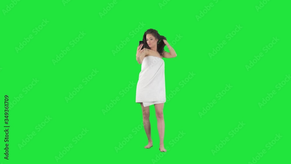 Young happy woman taking selfies with smart phone wrapped in bathroom cotton towel. Full body isolated on green screen background. 