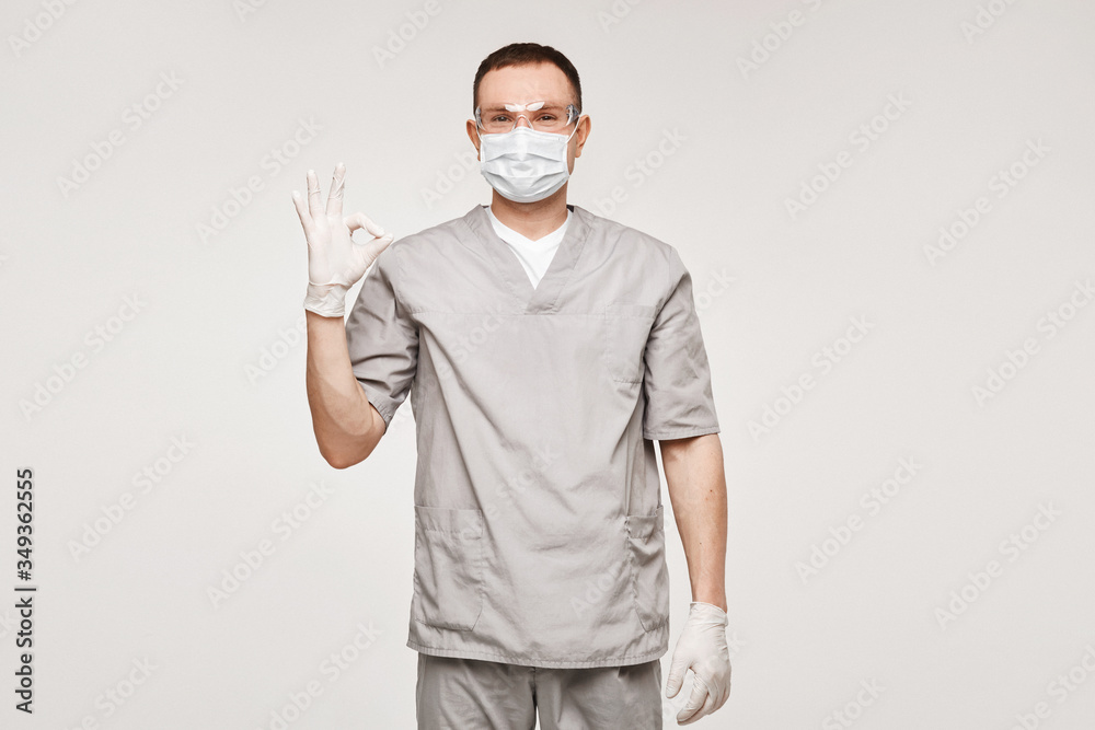 Young man in medical uniform, protective mask and glasses showing OK gesture with one hand and posing at the white background, isolated. Healthcare and emergency concept Young man in medical uniform, protective mask and glasses showing OK gesture with one hand and posing at the white background, isolated. Healthcare and emergency concept