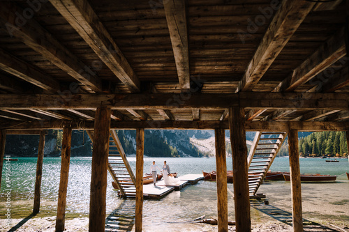 Fototapeta Naklejka Na Ścianę i Meble -  The bride and groom walk along a wooden boat dock at the Lago di Braies in Italy. Wedding in Europe, on Braies lake. Newlyweds walk, kiss, hug on a background of rocky mountains.