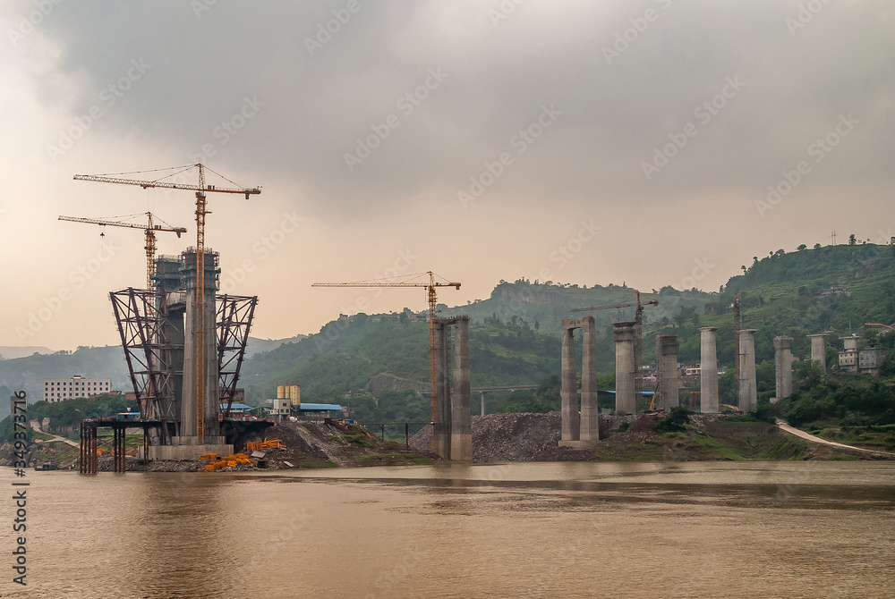 Fototapeta Fengdu, Chongqing, China - May 8, 2010: Yangtze River. Concrete suspension towers for bridge under construction with tall yellow cranes over brown water, cloudscape and green hill.