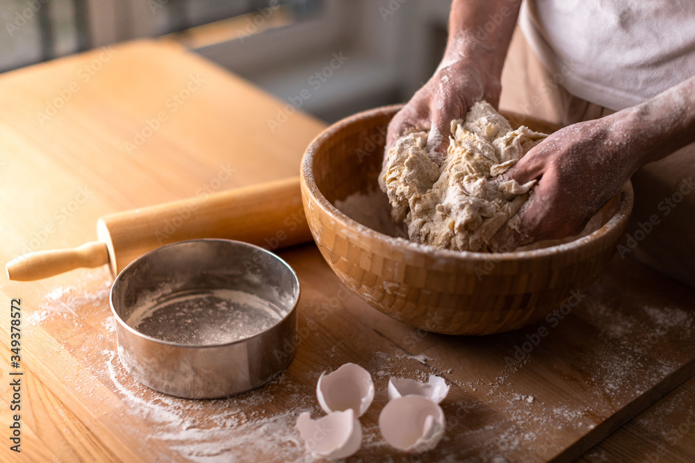 Cropped shot of man kneads dough for dumplings with his hands in a wooden bowl on the table. Eggshell, sieve and rolling pin by his side. 