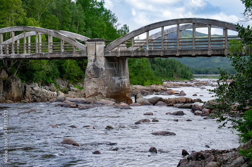 An old concrete archway bridge spans over a large salmon river with lots of rocks in the river.  There are tall green trees along both sides of the water. A hill or mountain is in the background.  photo