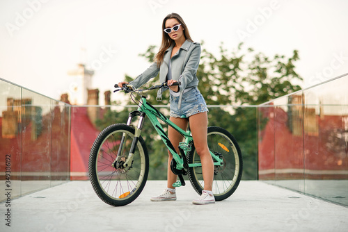 Attractive tempting young girl in fashion jeans clothes wears stylish glasses ,sitting on the bicycle and posing on camera at summer evening. Slow motion
