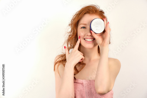 Redhead girl on a white background in a towel, laughing, covers one side of the face with a jar of cream.