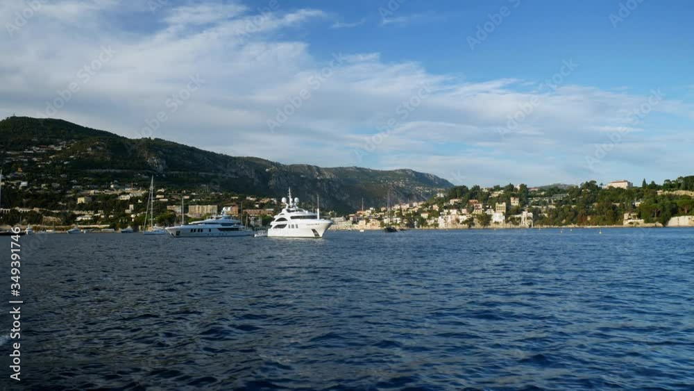 Luxury yachts on deep blue sea while leaving Nice port in France. French Riviera sailing on a sunny day to Monaco. Coastline in the background with mountain scenery. Rich lifestyle. Luxury yachts on deep blue sea while leaving Nice port in France. French Riviera sailing on a sunny day to Monaco. Coastline in the background with mountain scenery. Rich lifestyle.