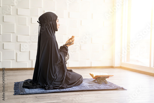beautiful asian muslim woman holding hands in chest embracing with prayer beads in her palm, praying to allah, facing down with hope and faith, wearing black hijab robe and bright cool warm background