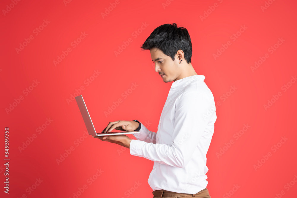 handsome asian businessman side view of him using laptop computer device hand on keyboard, wearing glasses and casual clothing and black earrings, standing and posing with red isolated background handsome asian businessman side view of him using laptop computer device hand on keyboard, wearing glasses and casual clothing and black earrings, standing and posing with red isolated background