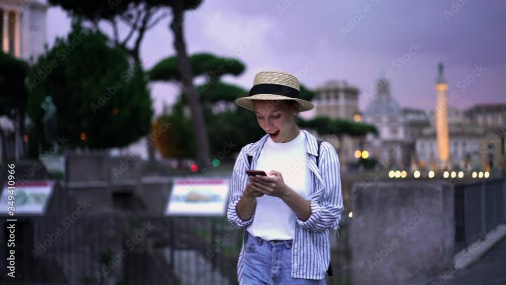 Slow motion of happy amazed young woman reading online news on website while having wonder reaction. Hipster girl received great news in email on smartphone walking on street in evening time
