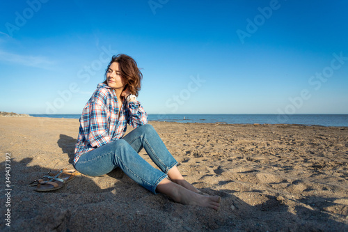 A cheerful dark-haired woman smiles, sitting on the beach,  enjoys the bright sun on a summer day. Concept of summer holidays at sea and live style © Виталий Сова