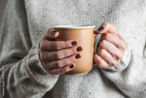 A woman wearing in a light cozy sweater is holding a cup of aromatic black coffee. The nails are coated with brown nail polish. Concept of coffee manicure.